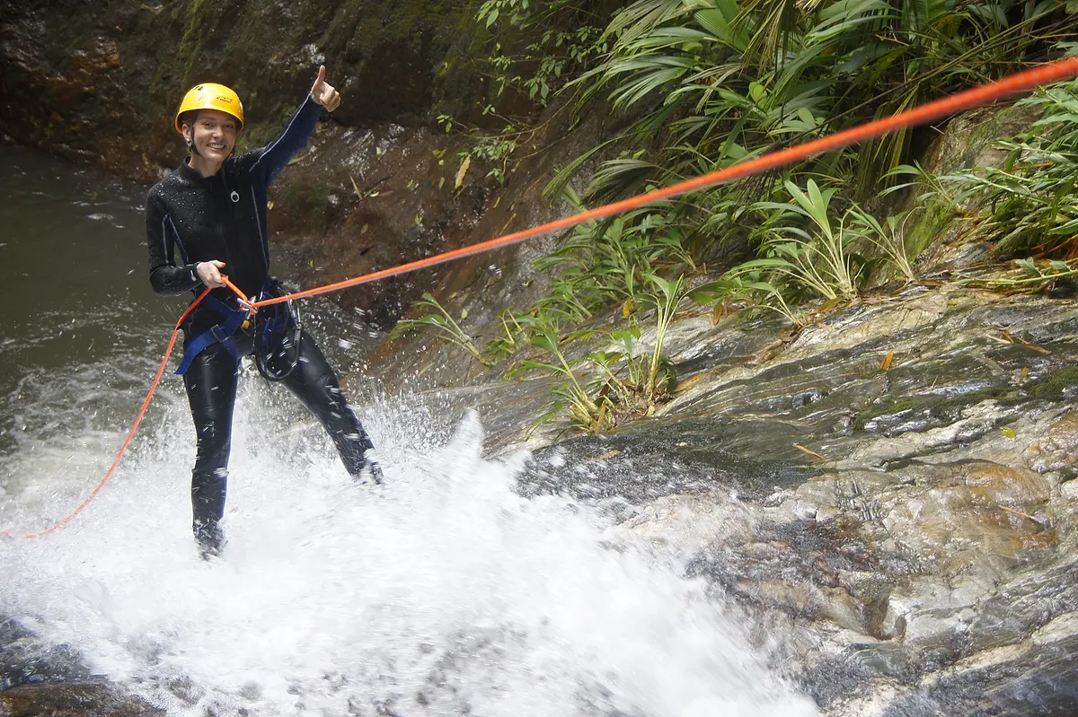 A smiling canyoner in a yellow helmet and wetsuit gives a thumbs-up while abseiling through white water on an orange rope