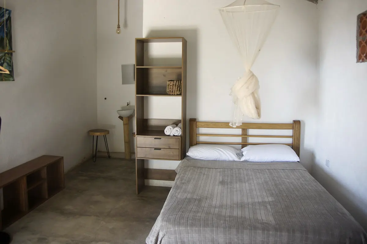 Deluxe double room: a wooden double bed with grey cover, a mosquito net knotted above, a tall wooden shelf and a low bench