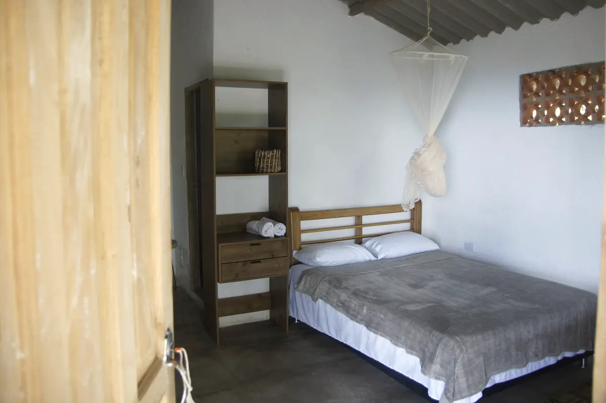 Deluxe double room seen from the doorway: double bed, mosquito net, wooden shelf and a patterned brick vent in the white wall
