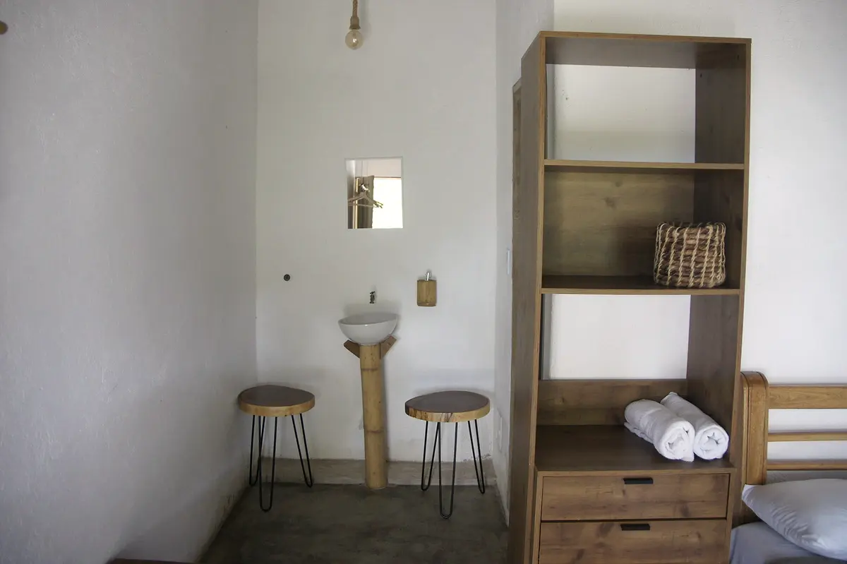 Corner of the deluxe double room with a bamboo-pedestal sink, two stools and a wooden shelf with rolled towels