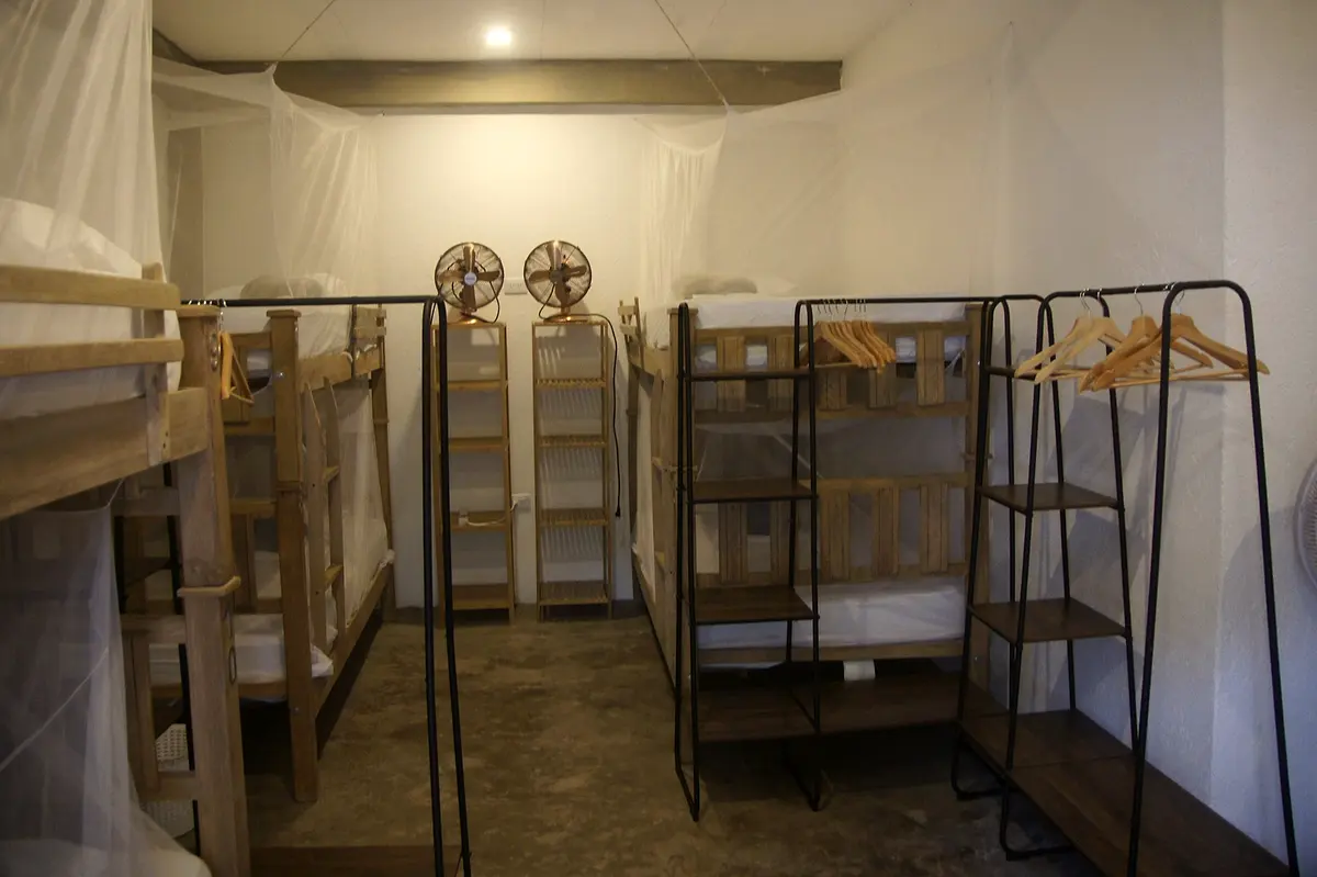 Wide view of the mixed dormitory with rows of wooden bunks, mosquito nets, wall fans and hanging rails