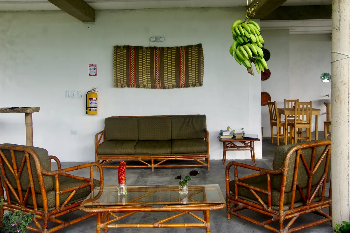 A rustic lounge with a green sofa, woven armchairs, a low table, a striped wall hanging and a bunch of bananas hanging from the beam