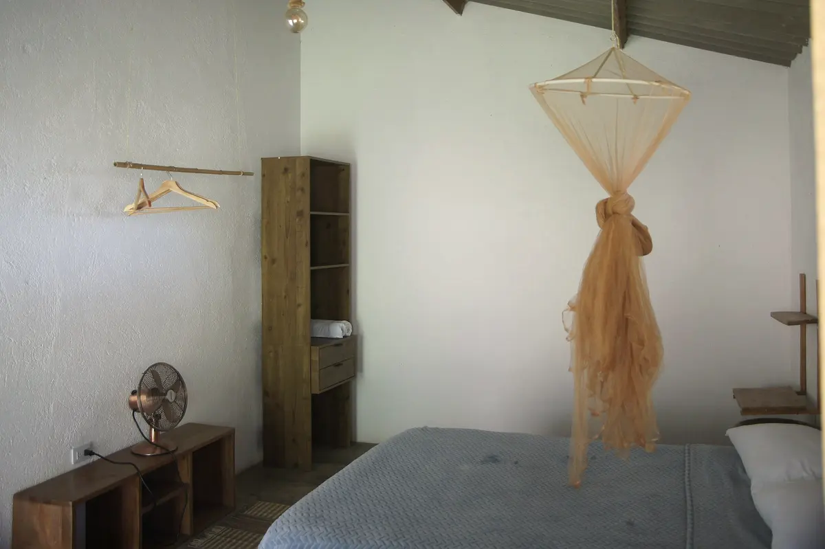 Double room with mountain view: double bed with grey cover, a knotted mosquito net, a desk fan, wooden hangers and a tall shelf