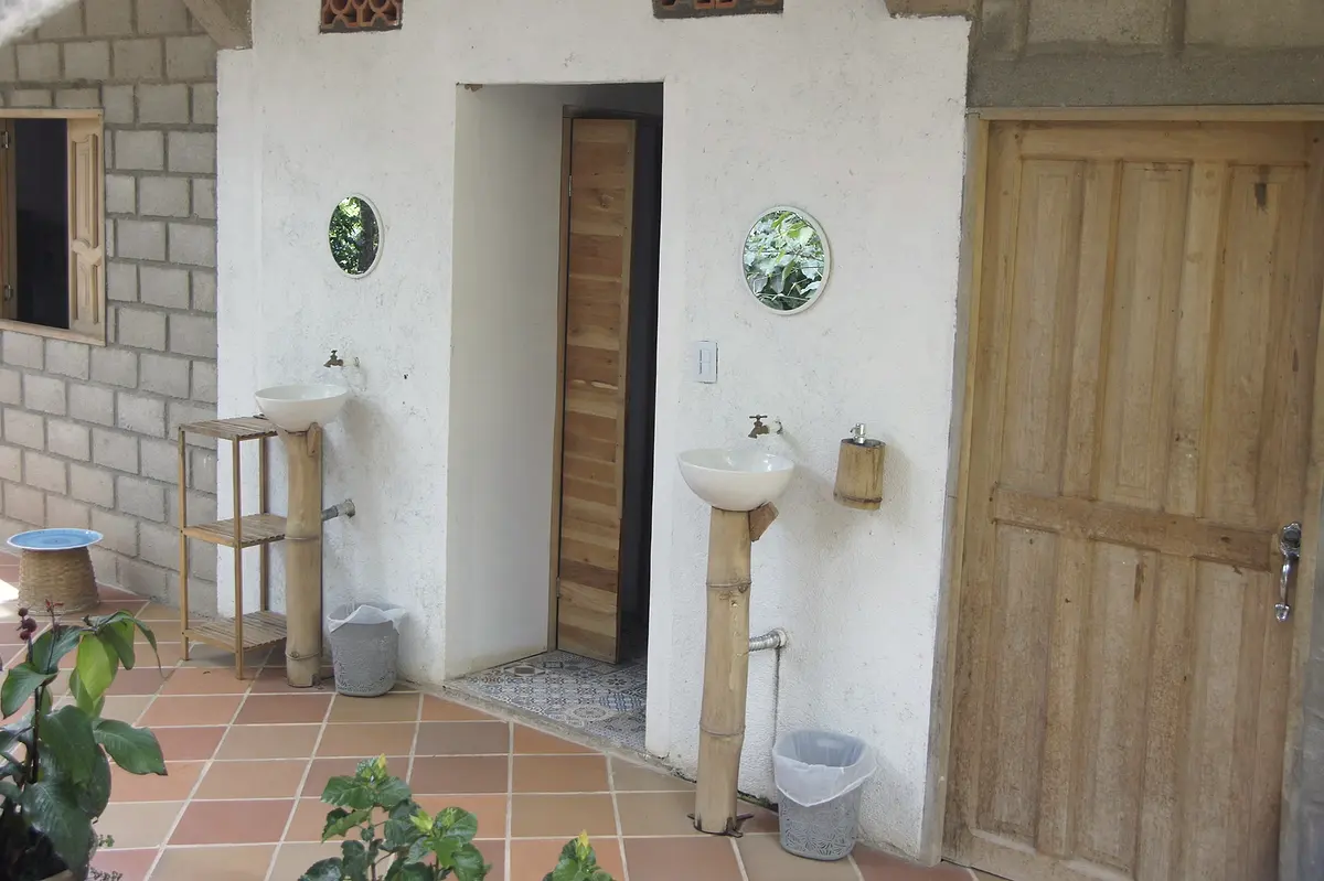 An open-air corridor with wooden doors, round mirrors and washbasins on bamboo pedestals beside a terracotta-tiled patio