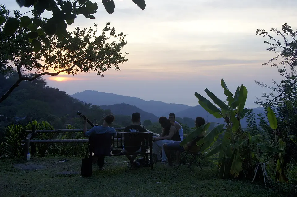 Guests sitting on a wooden bench in the garden at sunset, one holding a guitar, with banana plants and misty mountains beyond