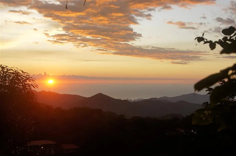 An orange sun dropping toward the sea behind dark ridges, framed by leaves, with hostel roofs in the foreground