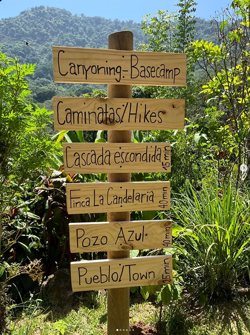 Hand-lettered wooden trail sign at the hostel: Canyoning-Basecamp, Caminatas/Hikes, Cascada escondida 15 min, Finca La Candelaria 40 min, Pozo Azul 40 min, Pueblo/Town 25 min